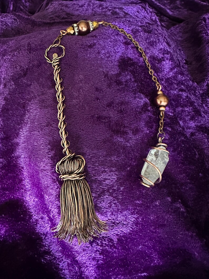 May include: A gold-toned pendulum with a clear quartz crystal and a tassel. The pendulum is hanging from a chain with brown beads and rhinestones.