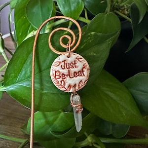 May include: A copper plant stake with a white ceramic charm that says "Just Be-Leaf" and a clear quartz crystal hanging from it.