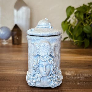 Peut inclure: Un pot cylindrique bleu clair avec un couvercle assorti, pr&eacute;sentant un relief sculpt&eacute; d'un visage entour&eacute; de cheveux tourbillonnants et d'un symbole de triple lune au-dessus. La surface a une finition mate textur&eacute;e avec de l&eacute;gers reflets blancs.