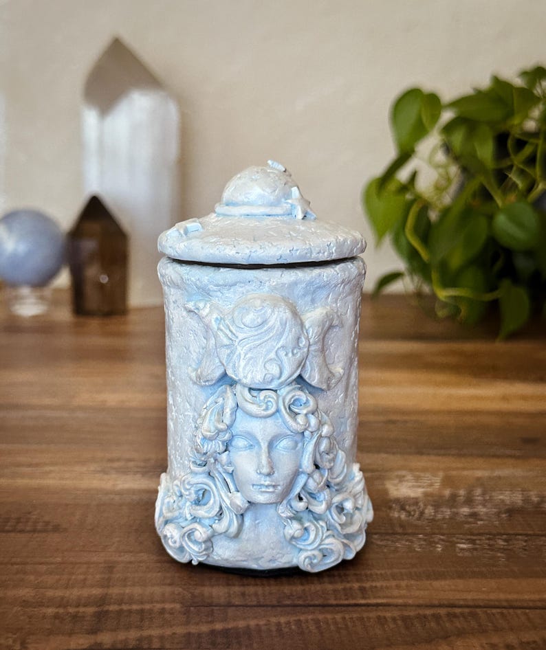 Peut inclure: Un pot cylindrique bleu clair avec un couvercle assorti, pr&eacute;sentant un relief sculpt&eacute; d'un visage entour&eacute; de cheveux tourbillonnants et d'un symbole de triple lune au-dessus. La surface a une finition mate textur&eacute;e avec de l&eacute;gers reflets blancs.