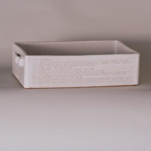Könnte beinhalten: Eine rechteckige, cremefarbene Keramik-Backform mit leicht strukturierter Oberfläche. Die Form hat an jeder Seite einen kleinen Griff und Text am oberen Rand. Der Text scheint ein Rezept oder Kochanweisungen zu sein.