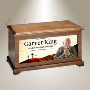 May include: A wooden cremation urn with a photo of a man and the text "Rest in Peace Garret King October 1940 - September 2018" on the front. The photo shows a man with white hair and a white beard, wearing a brown sweater and holding a walking stick. The background of the photo is a sunset with three crosses.