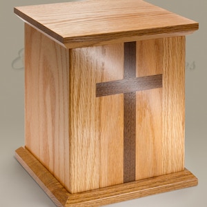 May include: A wooden cremation urn with a cross inlay. The urn is made of oak wood with a dark wood cross inlay. The urn has a square base and a rectangular body.