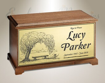 Puede incluir: Una urna de cremación de madera con una placa de latón. La placa presenta un dibujo de un sauce llorón sobre un banco junto a un lago. El texto en la placa dice "Rest in Peace Lucy Parker September 1947 - June 2018".