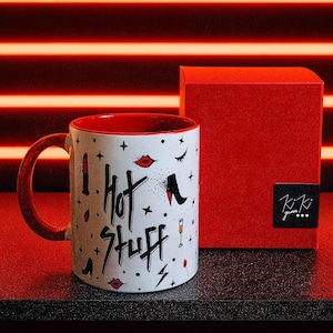 Op de afbeelding: Een witte keramische mok met een rode binnenkant en handvat, versierd met de tekst "Hot Stuff" en schoonheidsgerelateerde afbeeldingen. Een rode geschenkdoos staat naast de mok. Het ontwerp van de mok bevat lippenstift, hoge hakken en andere elementen.