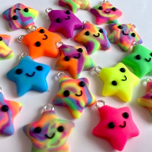 Op de afbeelding: Een verzameling kleurrijke ster-vormige bedels met lachende gezichten. De bedels zijn in verschillende kleuren, waaronder blauw, oranje, roze, paars, groen en geel, met een regenboog-wervelpatroon. Elke bedel heeft een zilveren lus.