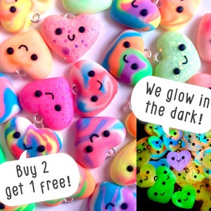 May include: A collection of colorful heart-shaped charms with cute faces. The charms are made of polymer clay and have a swirl pattern. Some of the charms are glittery. The charms glow in the dark. The text "Buy 2 get 1 free!" and "We glow in the dark!" is visible in the image.