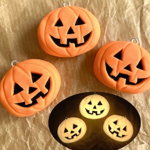 May include: Three orange Halloween jack-o'-lantern ornaments with black painted faces. The ornaments are hanging from silver strings. The ornaments are on a brown paper background.
