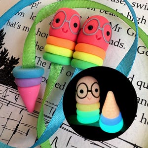 Può includere: Due segnalibri stravaganti in argilla polimerica fatti a mano. Presentano personaggi sorridenti a forma di verme rosso con occhiali, impilati su strati arcobaleno. Un'estremità del segnalibro abbinata e un nastro blu e verde completano il design. L'immagine include anche una versione luminosa.