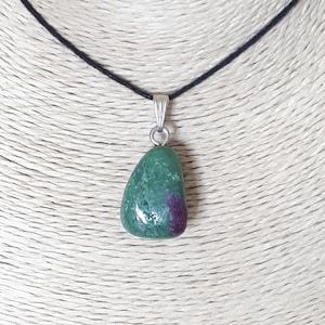 Może przedstawiać: Naszyjnik z wisiorkiem z wypolerowanym kamieniem szlachetnym w kształcie łzy. Kamień jest głównie zielony z plamą głębokiego czerwono-fioletowego koloru. Wisi na czarnym sznurku i ma srebrną metalową krawędź.