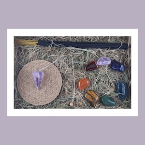 Peut inclure: Un assortiment d'articles spirituels dans une boîte, comprenant des bâtonnets d'encens, un pendule avec un cristal violet, un dessous de verre en liège avec un motif de fleur de vie et plusieurs pierres précieuses colorées. Les pierres sont de formes et de couleurs variées.