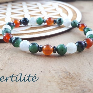 May include: A beaded bracelet featuring a variety of colorful gemstones, including white, green, orange, and black beads. The bracelet is displayed on a wooden surface. The word "Fertilité" is written in a teal script at the bottom.