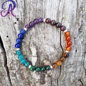 Peut inclure: Un bracelet de perles colorées présentant un spectre de couleurs, notamment le violet, le bleu, le vert, l'orange et le marron. Le bracelet est composé de pierres rondes et polies, assemblées avec des entretoises argentées. Il est présenté sur une surface en bois vieilli.
