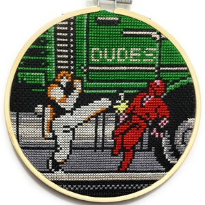 May include: A cross-stitch embroidery hoop depicting a scene from the video game 'Double Dragon'. The characters are a red-clad ninja and a white-clad martial artist, fighting in front of a green building with the word 'DUCES' on it.