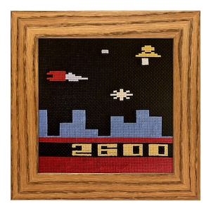 May include: A framed cross-stitch artwork depicting a pixelated video game scene. The scene features a spaceship, a UFO, and a cityscape with a score of 2600 displayed at the bottom.