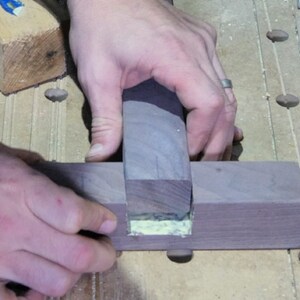 Puede incluir: Se muestran las manos de una persona sujetando una pieza de madera oscura, que se est&aacute; pegando en una ranura de otra pieza de madera oscura. La madera se mantiene en su lugar con un dedo.