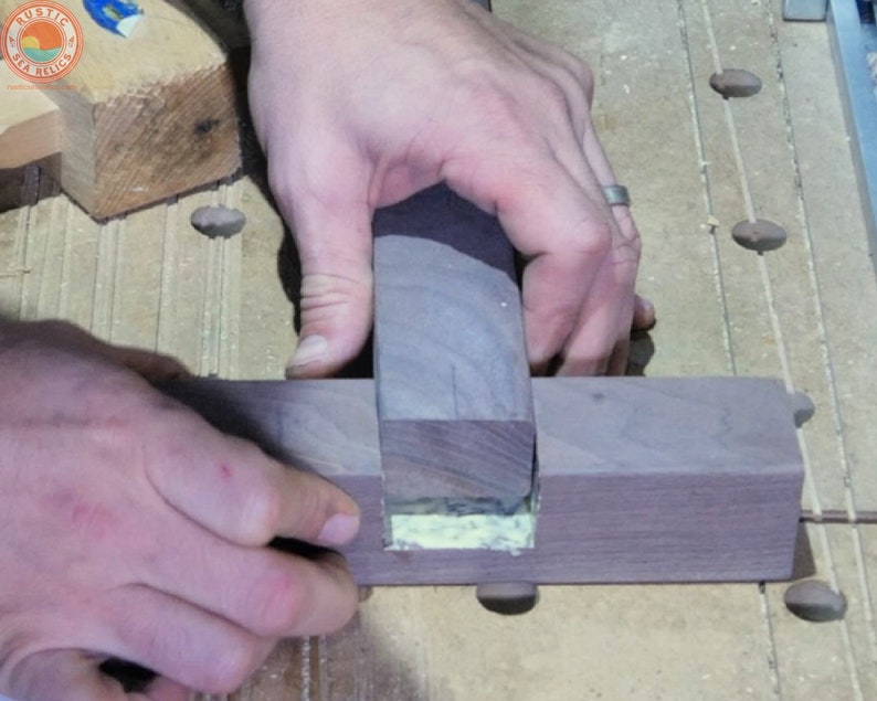 Puede incluir: Se muestran las manos de una persona sujetando una pieza de madera oscura, que se est&aacute; pegando en una ranura de otra pieza de madera oscura. La madera se mantiene en su lugar con un dedo.