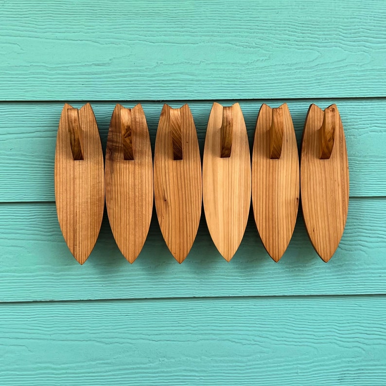 May include: Six wooden surfboard-shaped hooks in various shades of brown, mounted on a turquoise wall.