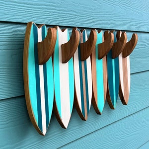 May include: A set of seven wooden surfboard-shaped hooks in turquoise and white with a natural wood finish. The hooks are mounted on a light blue wall.