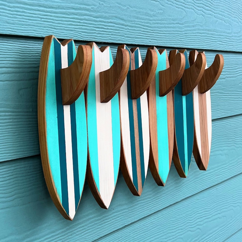 May include: A set of seven wooden surfboard-shaped hooks in turquoise and white with a natural wood finish. The hooks are mounted on a light blue wall.