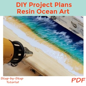 May include: A close-up of a resin ocean art project in progress. A heat gun is being used to cure the resin, which is poured over a wooden base. The resin is layered to create a realistic ocean scene with blue water, white waves, and a sandy beach.