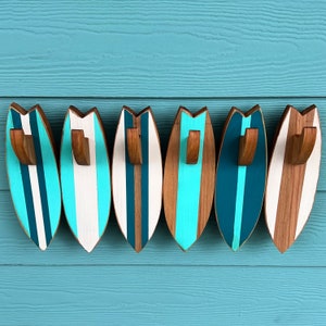 May include: Six wooden surfboard-shaped hooks with blue and white stripes. The hooks are mounted on a light blue wall.