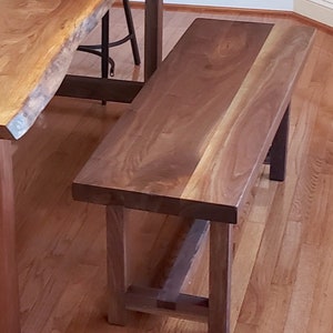 Puede incluir: Un banco de madera marr&oacute;n oscuro con un dise&ntilde;o sencillo. El banco tiene una parte superior rectangular y una base con dos patas a cada lado. El banco est&aacute; hecho de madera maciza y tiene un acabado natural.