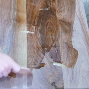 Puede incluir: Primer plano de una superficie de madera que se est&aacute; cubriendo con una resina epoxi transparente. La resina se est&aacute; extendiendo con una herramienta de madera. La madera tiene un patr&oacute;n de veta natural y es de color marr&oacute;n claro.