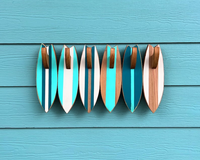 May include: Six wooden surfboard-shaped hooks in various shades of blue and brown, mounted on a light blue wall. The hooks are designed to resemble surfboards with a striped pattern.