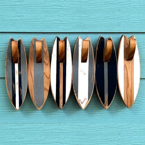 May include: Five wooden surfboard-shaped hooks with a black, white, and gray striped design. The hooks are mounted on a light blue wall.