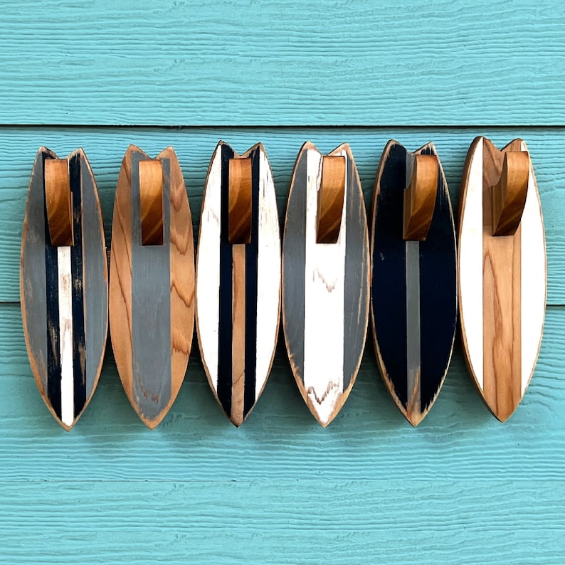May include: Five wooden surfboard-shaped hooks with a black, white, and gray striped design. The hooks are mounted on a light blue wall.