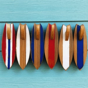 May include: Six wooden surfboard-shaped wall hooks in red, white, and blue. The hooks are painted with stripes and have a rustic, beachy feel.