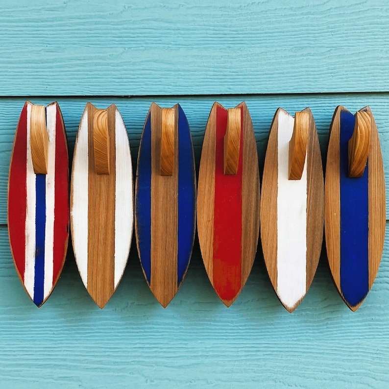 May include: Six wooden surfboard-shaped wall hooks in red, white, and blue. The hooks are painted with stripes and have a rustic, beachy feel.