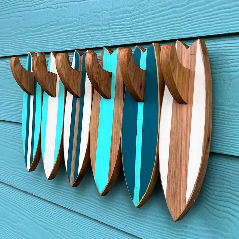 May include: A set of seven wooden surfboard wall decor in various shades of blue and white. The surfboards are arranged in a row, with the largest surfboard on the right and the smallest on the left. The surfboards are all painted with a simple design of stripes.