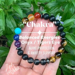 May include: A black beaded bracelet with seven colorful chakra stones, onyx, and tiger's eye beads. The bracelet has the text "Chakra" and "Balanced Energies Onyx / Tigers Eye Auric Protection & Anxiety Support" printed on it. The text "SPHINX CRYSTAL THERAPY" is also printed on the bracelet.