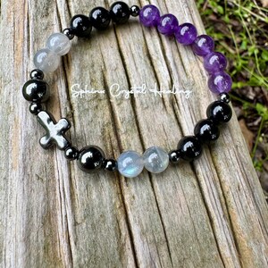 May include: A beaded bracelet featuring round black, purple, and iridescent gray beads, interspersed with small metallic spacers and a metallic cross charm. The text "Sphinx Crystal Healing" is printed in white across the center of the bracelet.