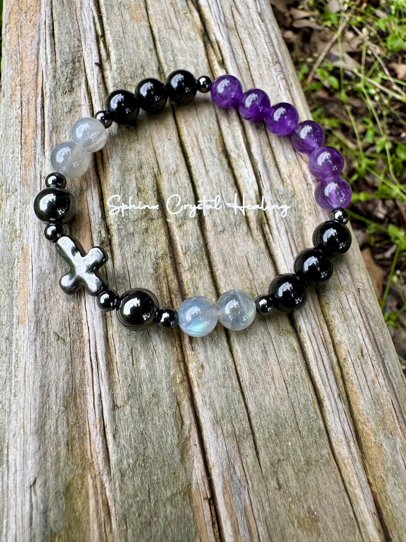 May include: A beaded bracelet featuring round black, purple, and iridescent gray beads, interspersed with small metallic spacers and a metallic cross charm. The text "Sphinx Crystal Healing" is printed in white across the center of the bracelet.