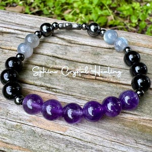 May include: A beaded bracelet featuring a sequence of polished purple amethyst, glossy black obsidian, translucent grey, and metallic hematite beads. The text Sphinx Crystal Healing is overlaid in white script across the center of the bracelet.