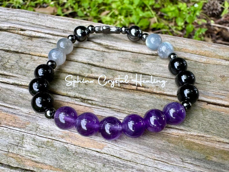 May include: A beaded bracelet featuring a sequence of polished purple amethyst, glossy black obsidian, translucent grey, and metallic hematite beads. The text Sphinx Crystal Healing is overlaid in white script across the center of the bracelet.