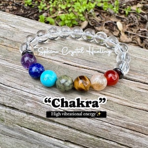 May include: A clear crystal bracelet with seven chakra stones: amethyst, lapis lazuli, turquoise, aventurine, tiger's eye, carnelian, and red jasper. The bracelet is on a wooden surface and the text "Chakra" and "High vibrational energy" is below the bracelet.