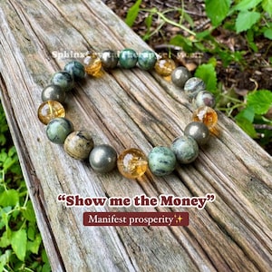 May include: A beaded bracelet featuring alternating round polished stones in shades of green, grey, and translucent yellow. The image includes the text "Show me the Money" and "Manifest prosperity" with two small star icons printed on the surface.