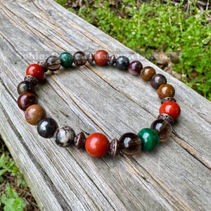 May include: A beaded bracelet featuring a variety of colorful gemstones, including red, green, brown, and black beads. The bracelet is resting on a weathered wooden surface, showcasing its natural tones and textures. The beads are approximately 0.5 inches in diameter.