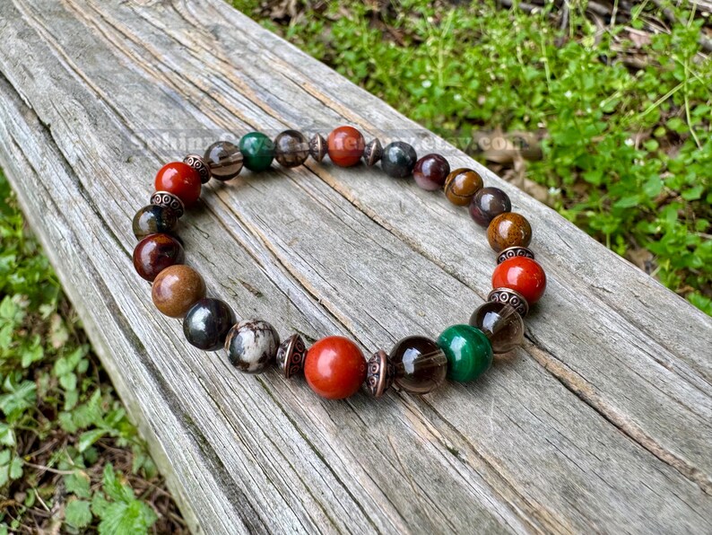 May include: A beaded bracelet featuring a variety of colorful gemstones, including red, green, brown, and black beads. The bracelet is resting on a weathered wooden surface, showcasing its natural tones and textures. The beads are approximately 0.5 inches in diameter.
