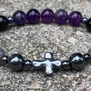 May include: A beaded stretch bracelet featuring a central metallic cross charm, flanked by dark gray hematite beads, smooth purple amethyst spheres, glossy black beads, and iridescent gray labradorite stones, all arranged on a circular elastic cord.