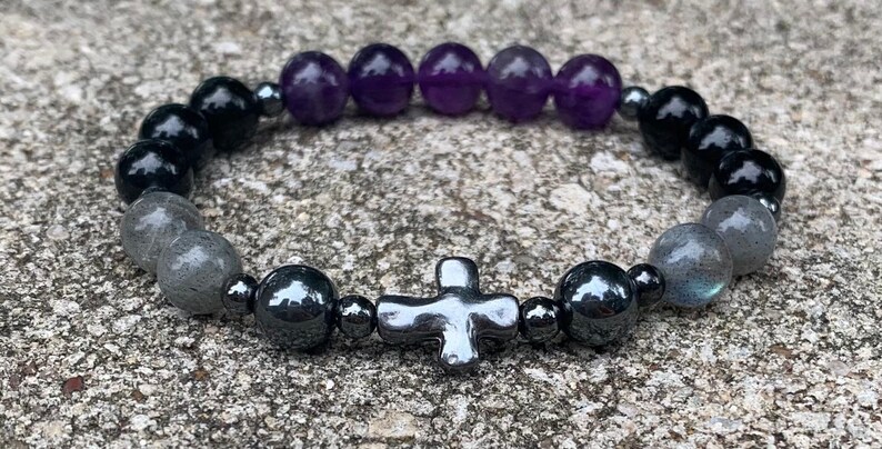 May include: A beaded stretch bracelet featuring a central metallic cross charm, flanked by dark gray hematite beads, smooth purple amethyst spheres, glossy black beads, and iridescent gray labradorite stones, all arranged on a circular elastic cord.