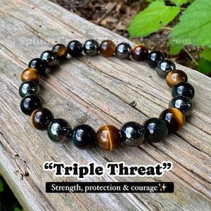 May include: A beaded bracelet featuring alternating smooth, polished spherical beads in shades of golden brown tiger's eye, deep black obsidian, and metallic hematite. The text "Triple Threat" and "Strength, protection & courage" is visible.