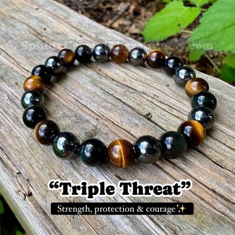 May include: A beaded bracelet featuring alternating smooth, polished spherical beads in shades of golden brown tiger's eye, deep black obsidian, and metallic hematite. The text "Triple Threat" and "Strength, protection & courage" is visible.