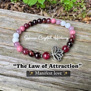 May include: A beaded bracelet with a variety of pink, red, and white stones. The bracelet has a silver lotus charm and the text "The Law of Attraction" and "Manifest love". The bracelet is on a wooden surface.