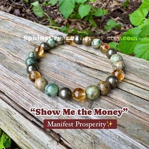 May include: A beaded bracelet featuring alternating round polished stones in shades of green, brown, and translucent yellow. The text "Show Me the Money" and "Manifest Prosperity" is printed below the bracelet on the wooden surface.