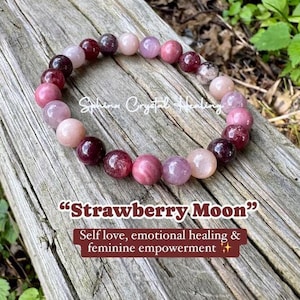 May include: A "Strawberry Moon" bracelet made of round, polished beads in shades of pink and burgundy. The bracelet is displayed on a weathered wooden surface. Text on the image reads "Self love, emotional healing & feminine empowerment."